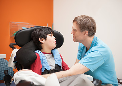 Father talking with disabled biracial son sitting in wheelchair while waiting in doctor's office, laughing together.